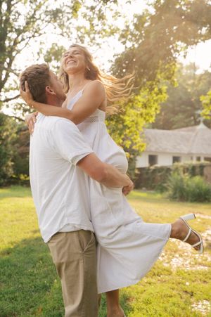 A couple embraces joyfully in a sunny meadow at golden hour, both wearing white casual attire.