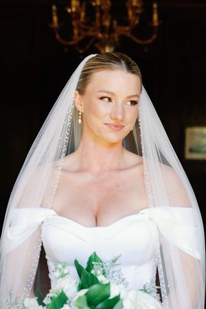A bridal portrait shows someone in an elegant off-shoulder white wedding dress with a flowing veil holding green foliage.