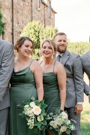 Wedding party in grey suits and emerald green dresses holding white floral bouquets stand together outside stone building.