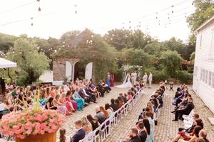 Wedding ceremony guests sit in rows of white chairs on a paved outdoor terrace overlooking gardens and trees.