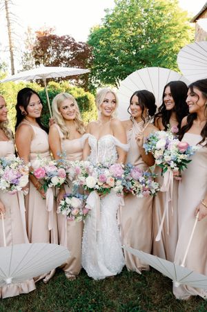 Bridal party wearing champagne-colored dresses stands in a line holding pink and white floral bouquets outdoors.