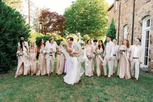 Wedding party in neutral beige and cream attire walks and poses on grassy lawn near historic stone building.