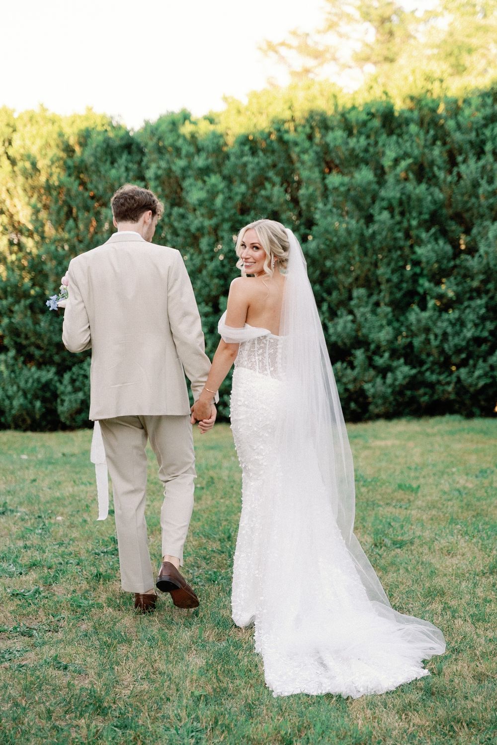 Newlywed couple walks hand in hand on a grassy lawn with lush green hedges in the background.