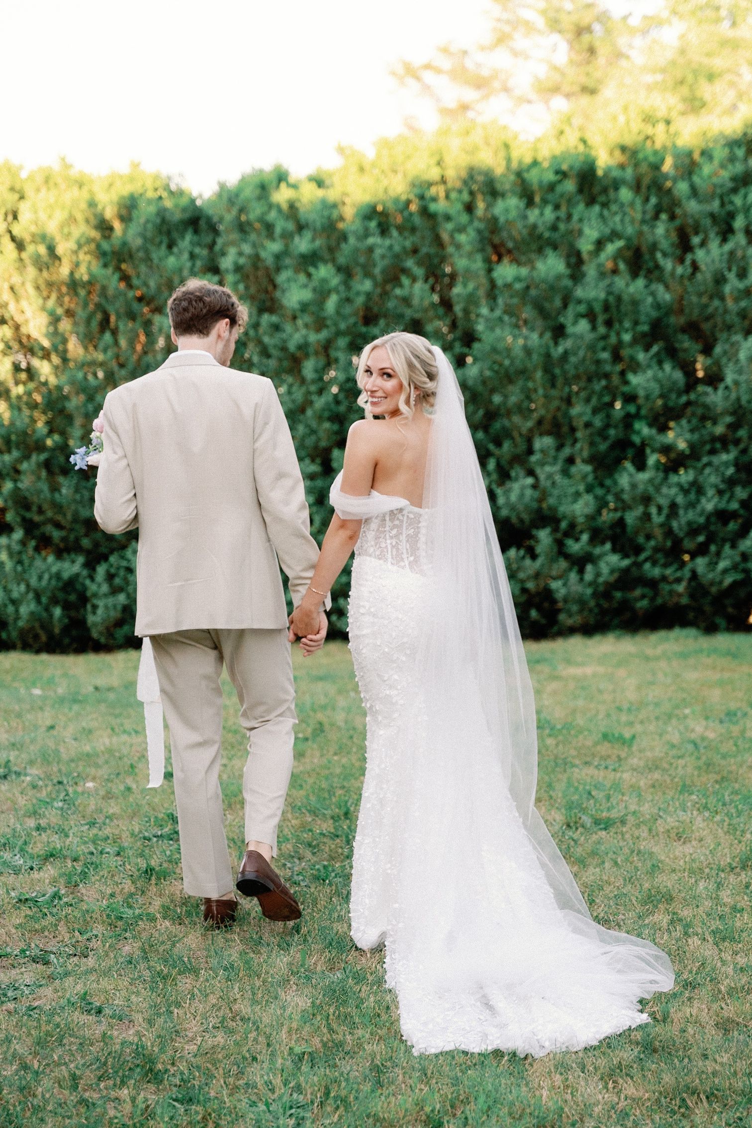 Newlywed couple walks hand in hand on a grassy lawn with lush green hedges in the background.