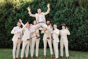 Wedding party in cream colored attire poses joyfully against lush green hedge background for celebratory photos.