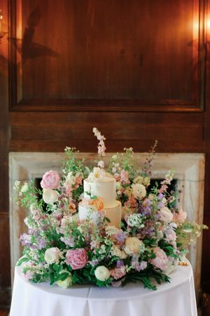 Elegant white wedding cake adorned with pastel floral arrangements of pink, purple, and white blooms on a white tablecloth.