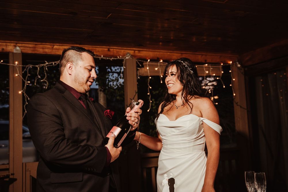 Bride smiling at groom lovingly as he opens a bottle of champagne after their elopement