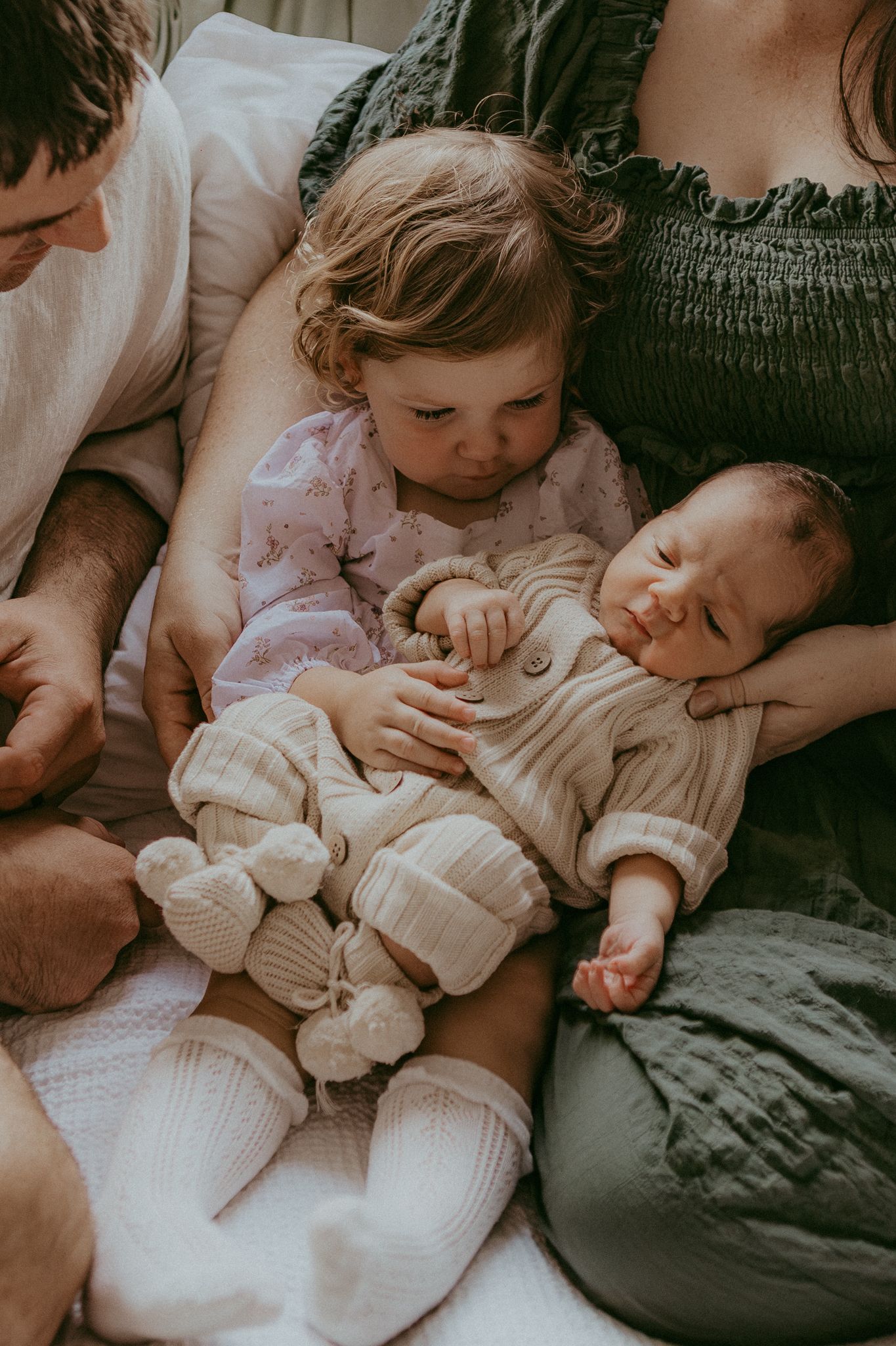 Toddler in pink pajamas gently holding a sleeping newborn baby while nestled between parents on a soft bed.