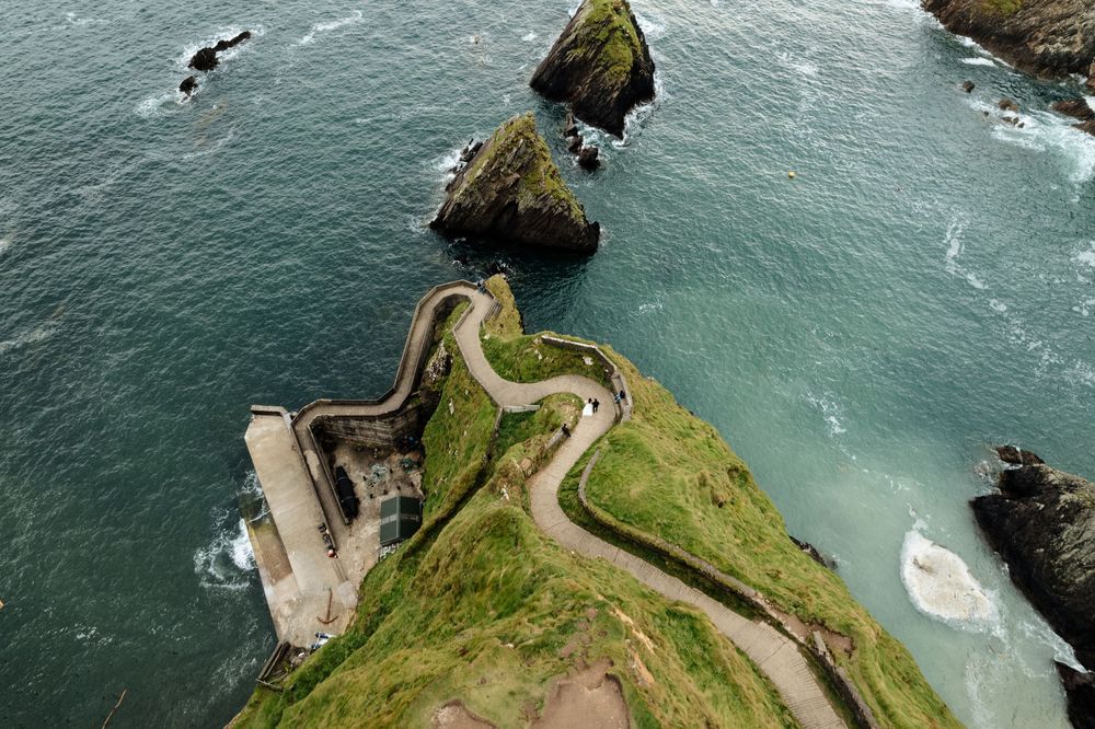 Aerial view of a winding coastal road leading to Dunquin Pier on Ireland's Dingle Peninsula with turquoise waters below.