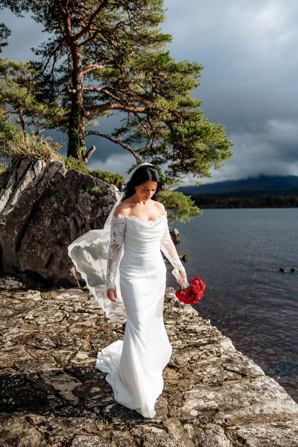 A bride in a white wedding dress stands on rocky shoreline with evergreen trees and stormy lake waters in the background.