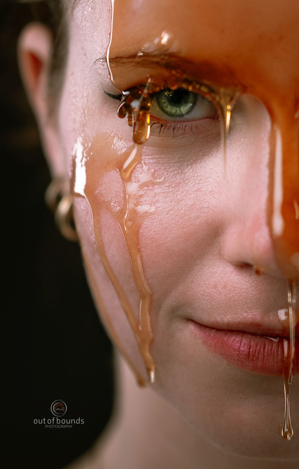 Artistic portrait with golden honey dripping down skin near a bright green eye against dark background.