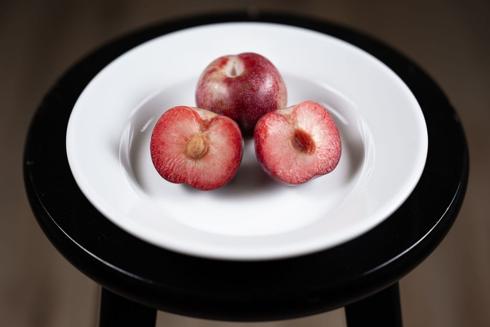 Three red plumbs arranged on a white plate atop a black circular table surface.