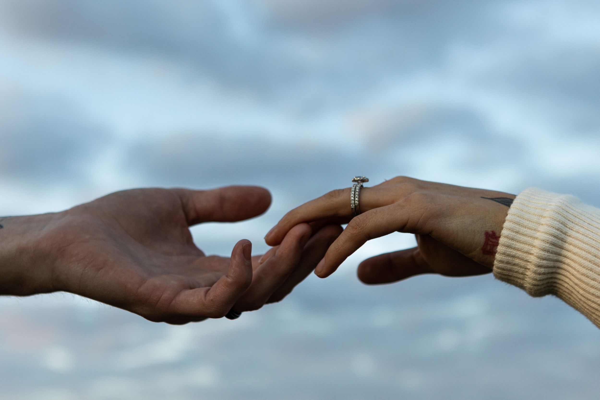 A newly engaged couple brushes hands with the sky as the background. You can see a few tattoos and an engagement ring