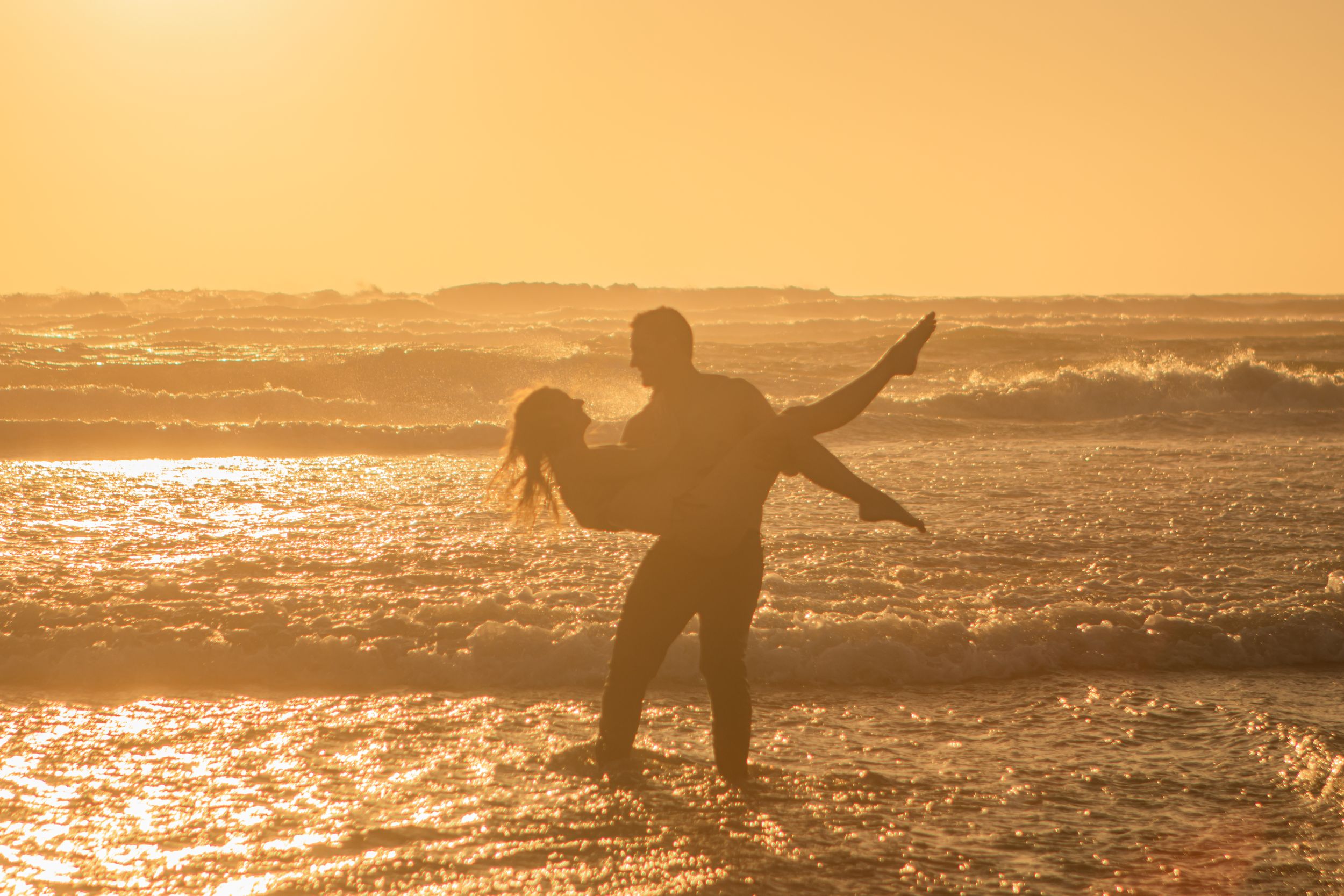 A couple dances in the ocean. There's romance in the air, and the sun is setting off to the side.