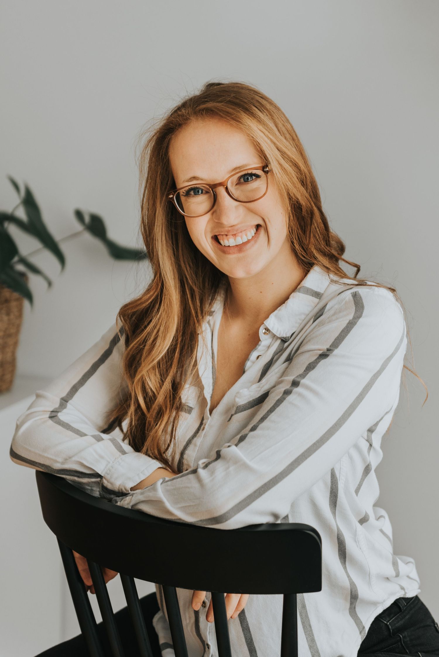 A professional portrait shows someone with glasses and long blonde hair wearing a white button-down shirt seated on a black chair.