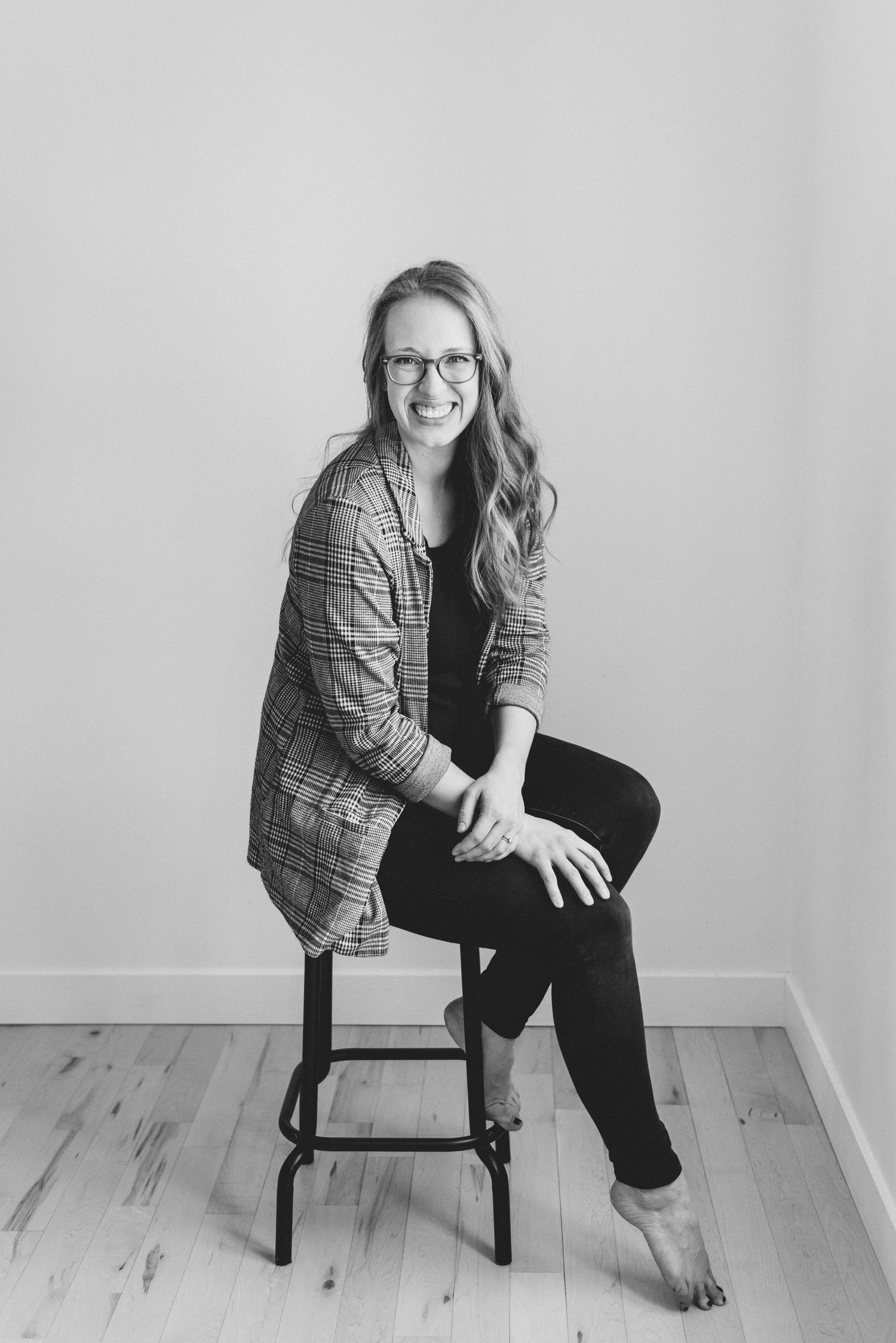 Black and white portrait of someone wearing glasses and plaid blazer sitting casually on wooden stool against light wall.