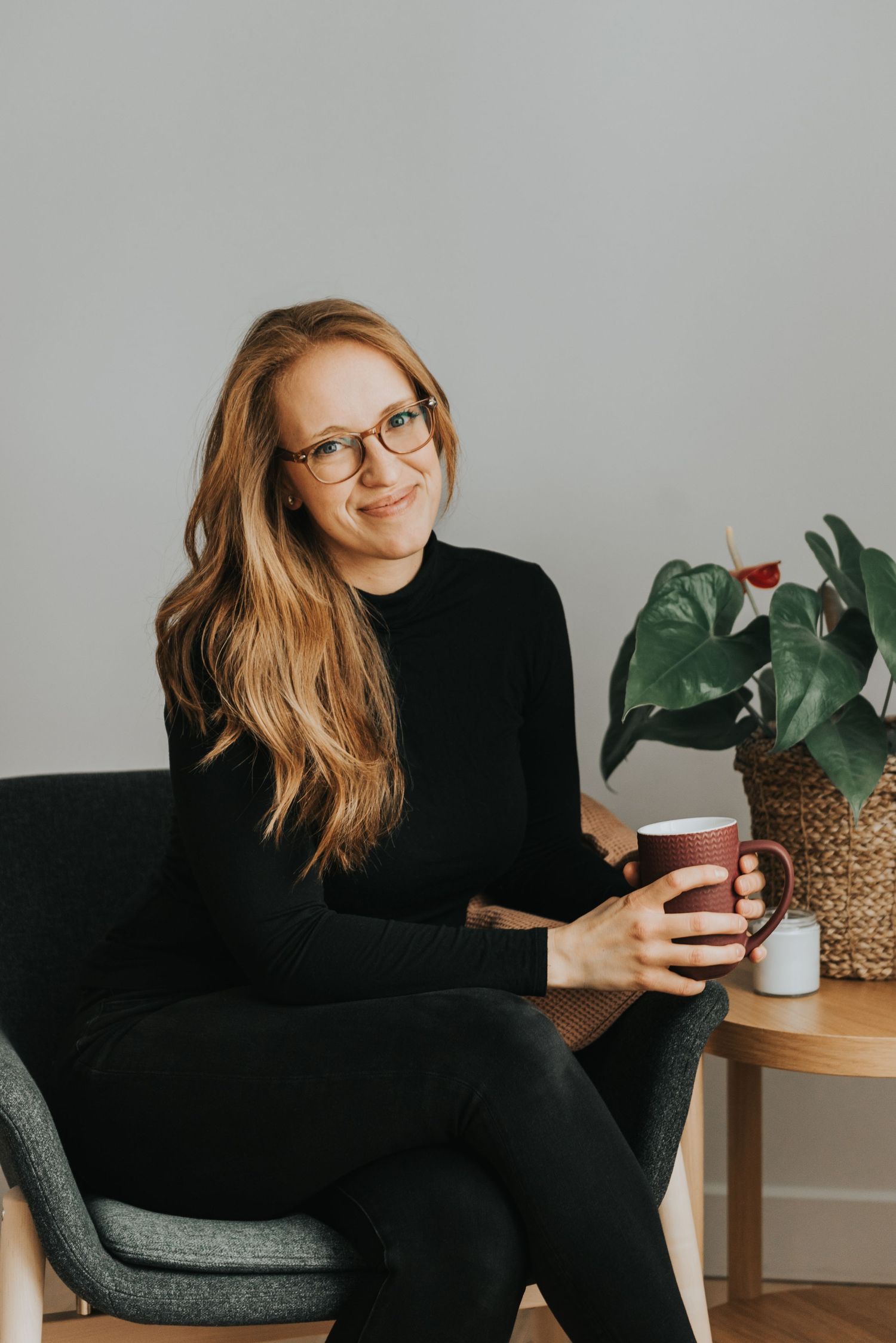 A person in a black sweater sits in a gray chair holding a coffee cup, with a potted plant on a side table nearby.