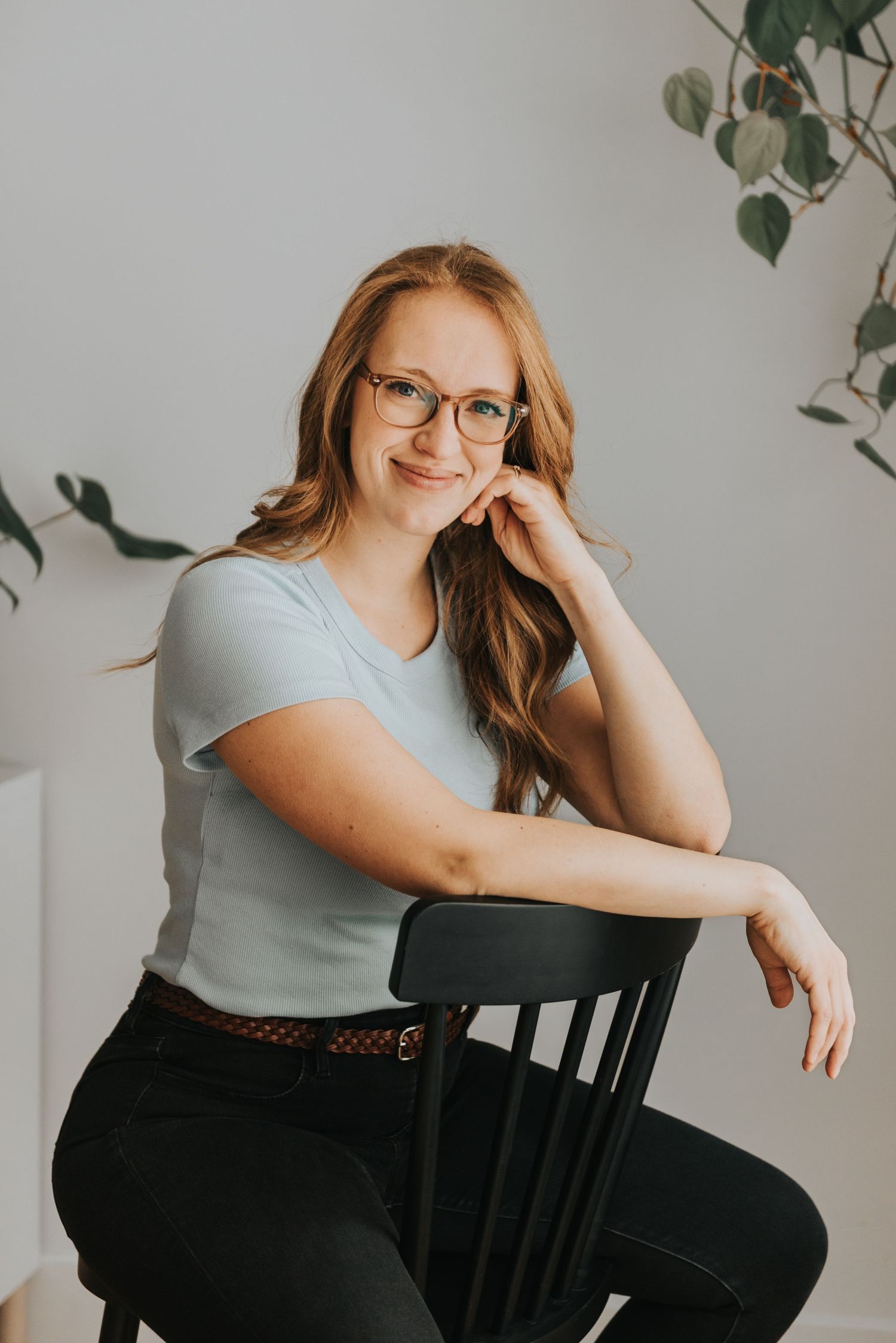 A person in glasses sits casually on a black wooden chair, wearing a light blue t-shirt against a white wall with plant decor.