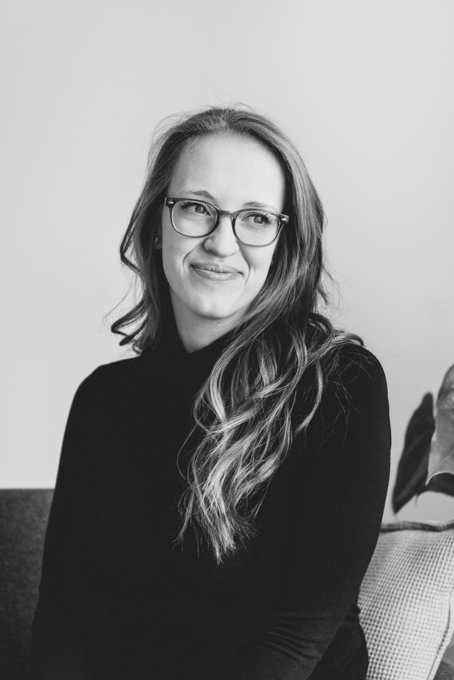 Black and white portrait of a smiling person with glasses and long wavy hair wearing a dark sweater.