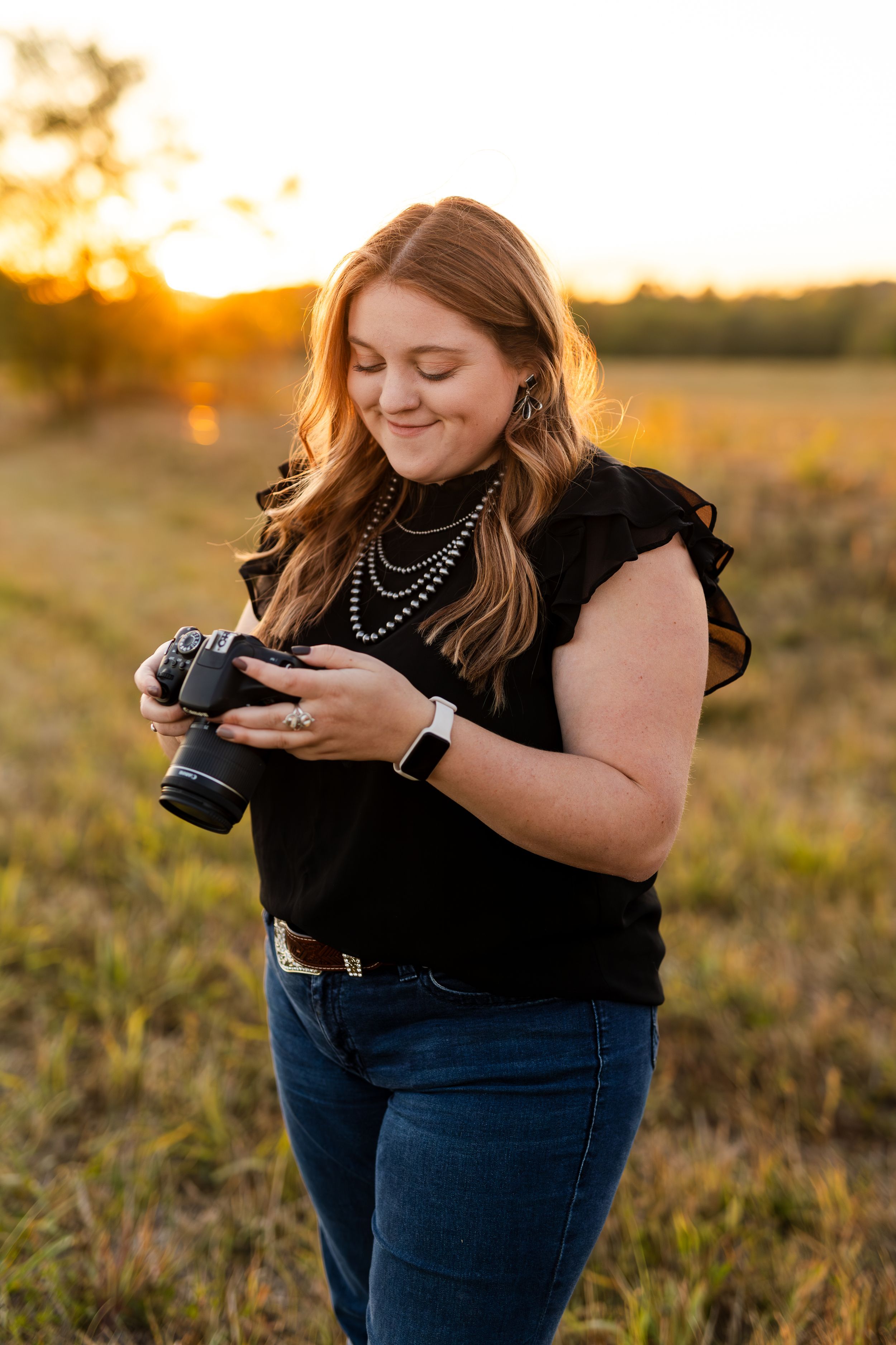 Woman in black top holding DSLR camera at golden hour in a field, wearing pearl necklace and jeans.