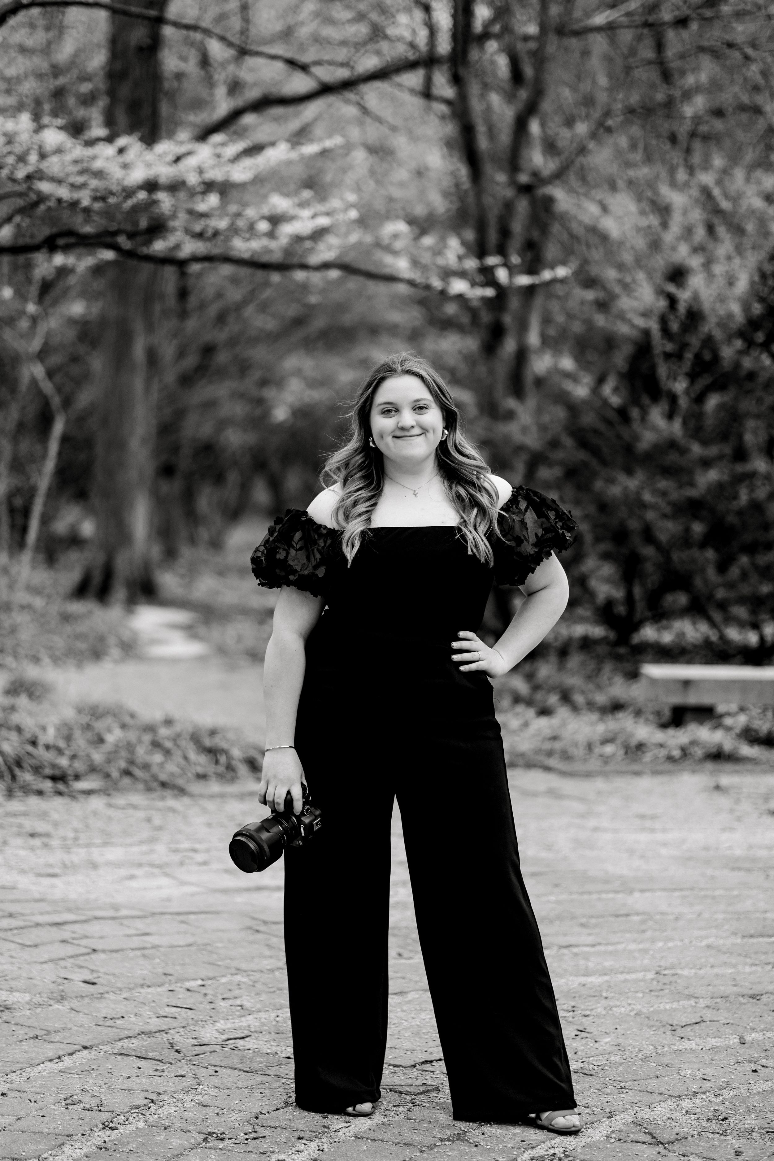 Laura in black jumpsuit holds a camera while posing on a brick path in a wooded park.