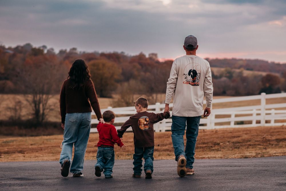 Family walking together on a rural road during autumn with white fence and fall foliage in background.