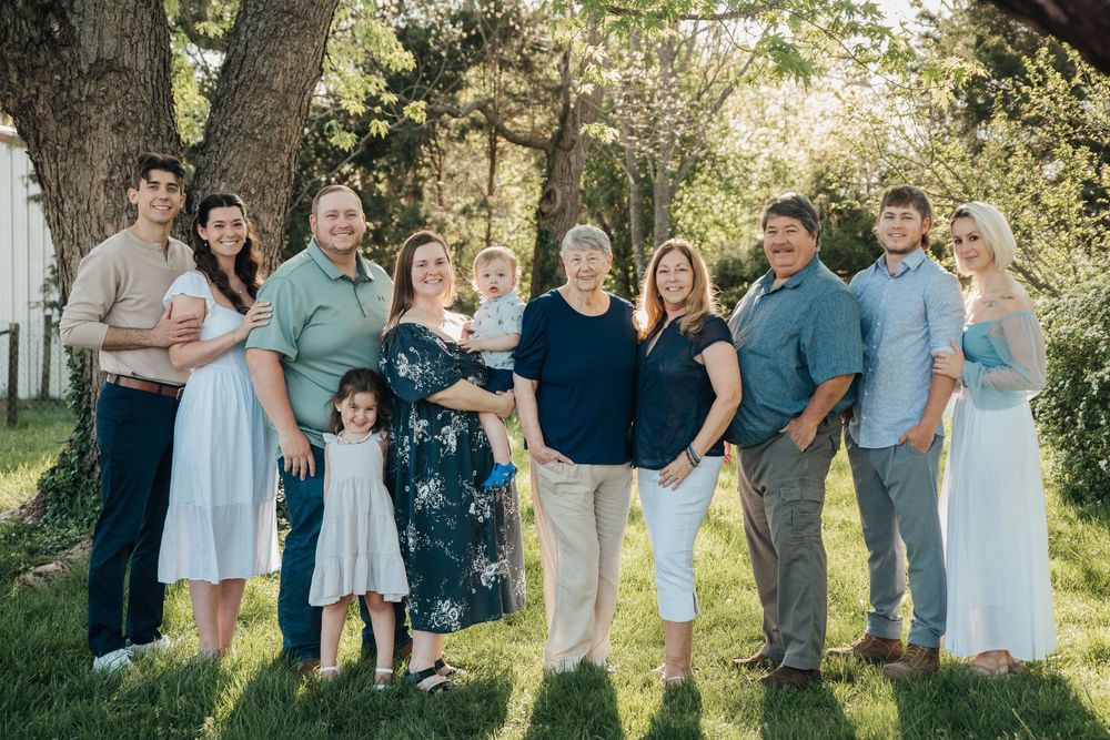 A large multi-generational family stands together outdoors under a tree on a sunny spring day.