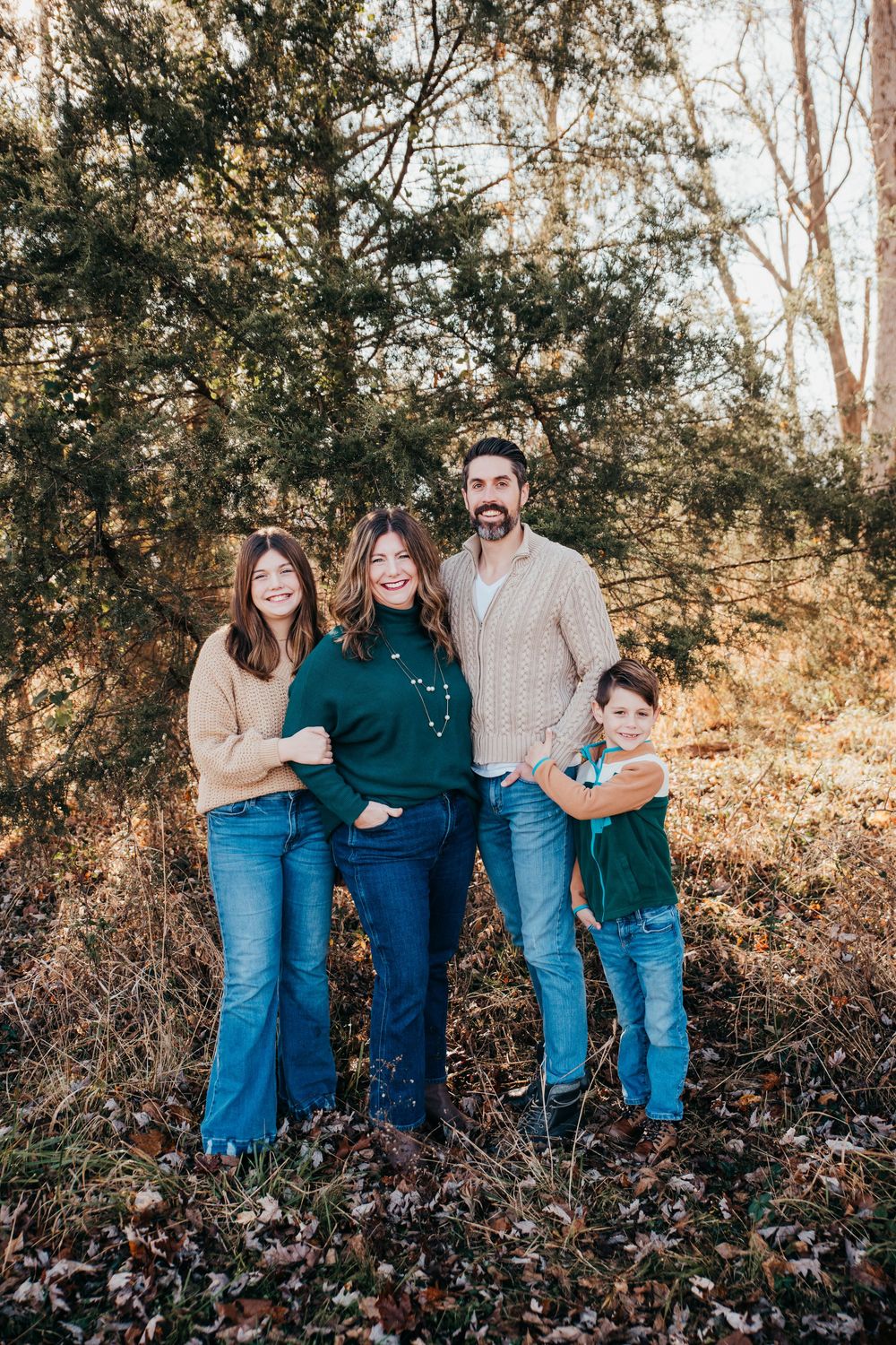 A family of four poses together outdoors on an autumn day wearing casual denim and sweaters in a wooded setting.