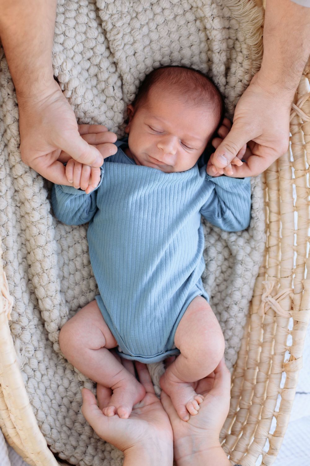A newborn baby wearing a light blue onesie rests on a textured knit blanket while gentle hands hold their arms.