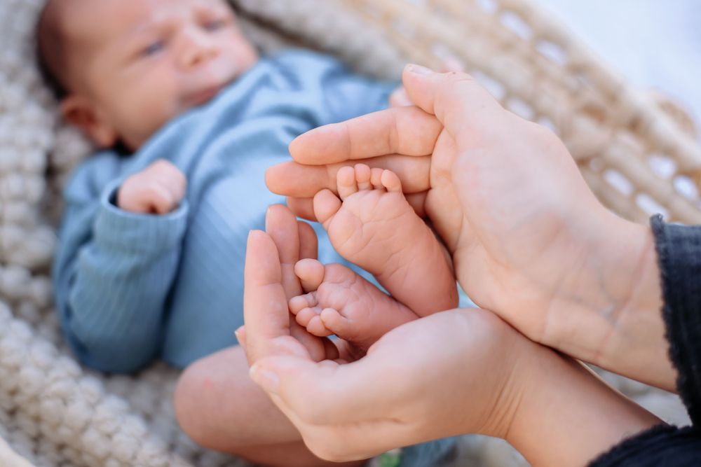 Caring hands cradle tiny feet of a baby in blue against a cozy knitted backdrop.