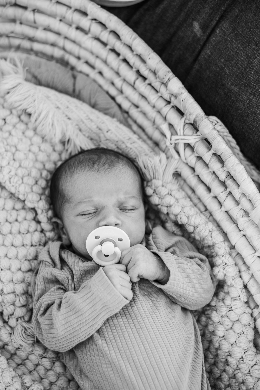 A sleeping infant with a pacifier lies peacefully in a woven basket with knit blanket.