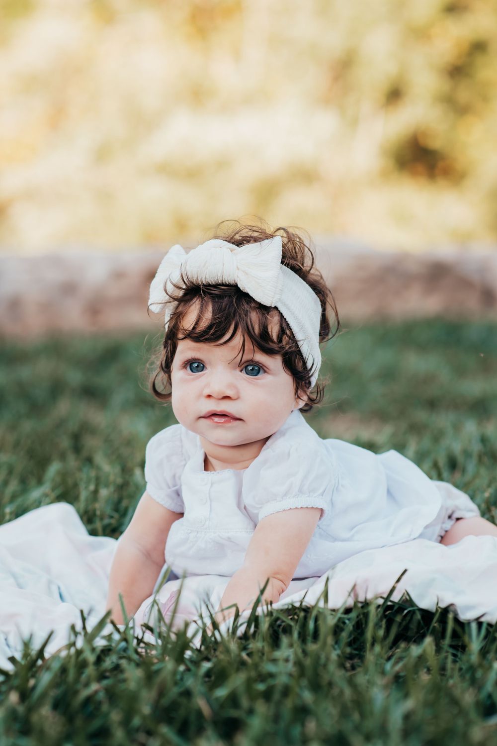 Baby in white dress and bow headband crawling on grass during outdoor photoshoot.