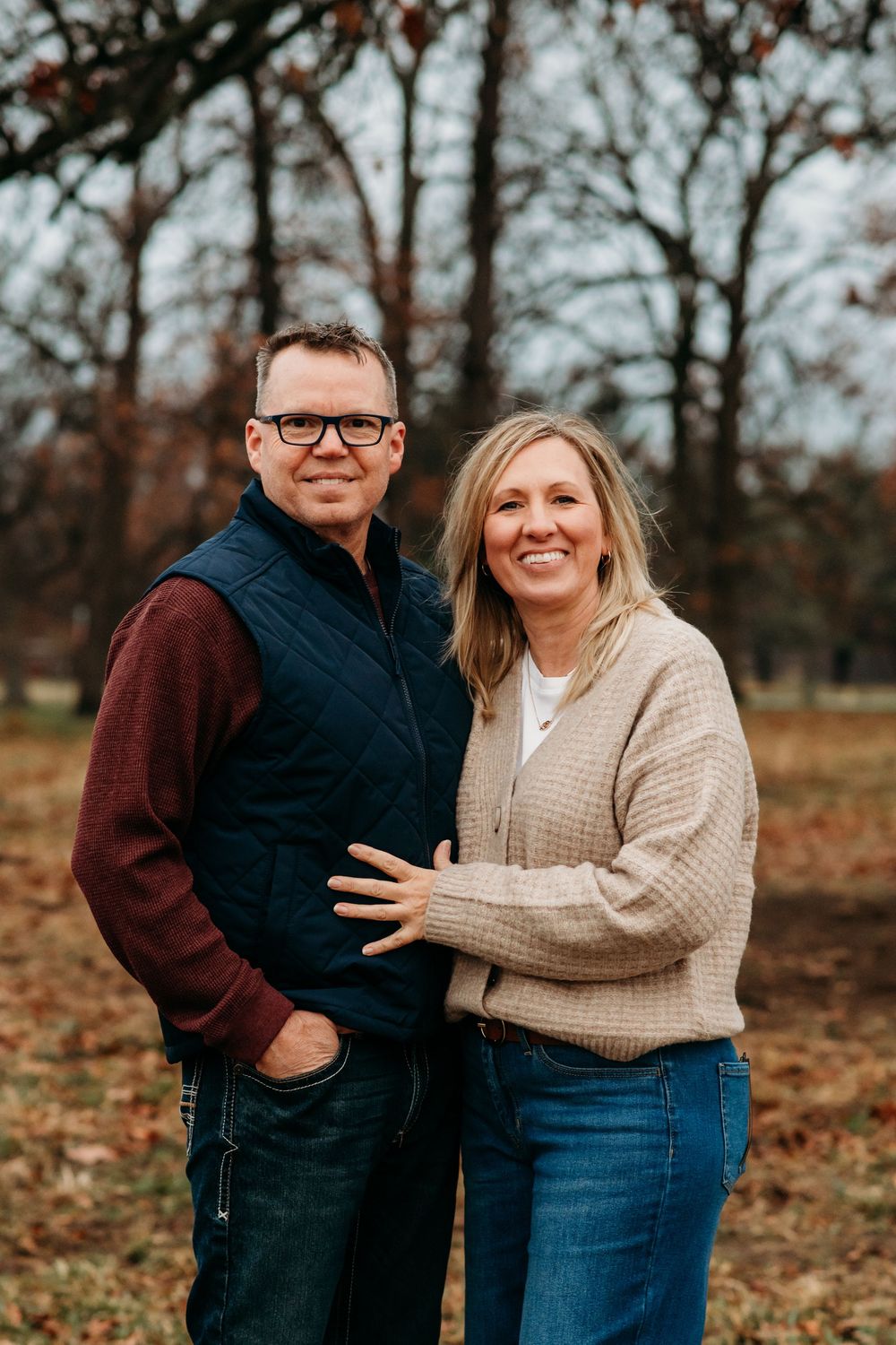 A couple in casual fall attire poses outdoors among autumn trees for a portrait in a natural setting.