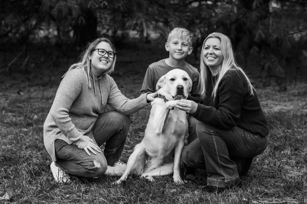 Black and white photo of two people outdoors sitting on grass with a friendly Labrador dog between them.