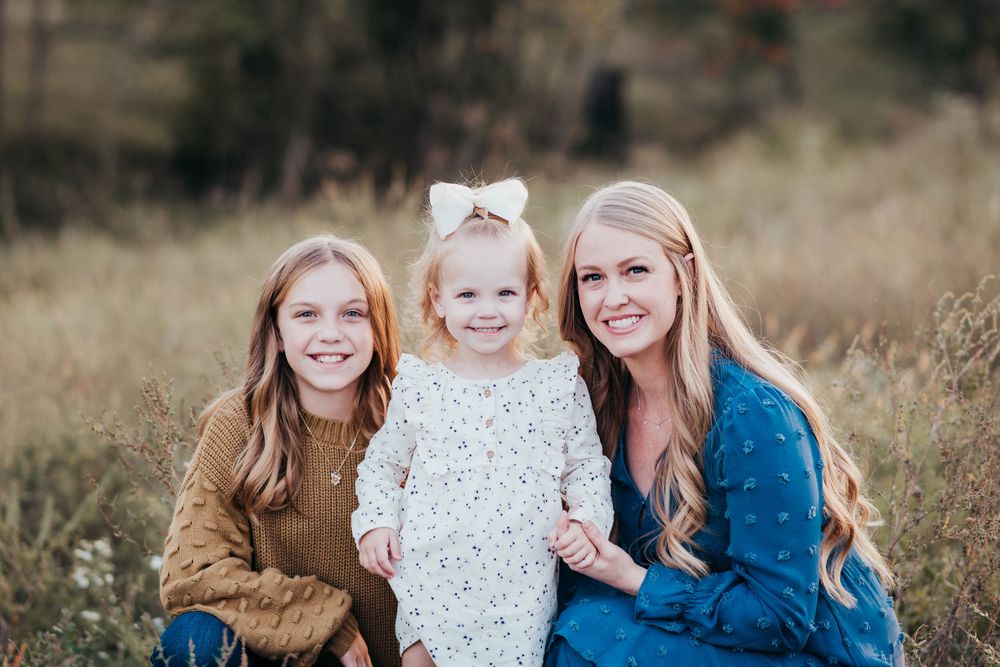 Three people smiling together in a field with tall grass at golden hour wearing casual fall attire.