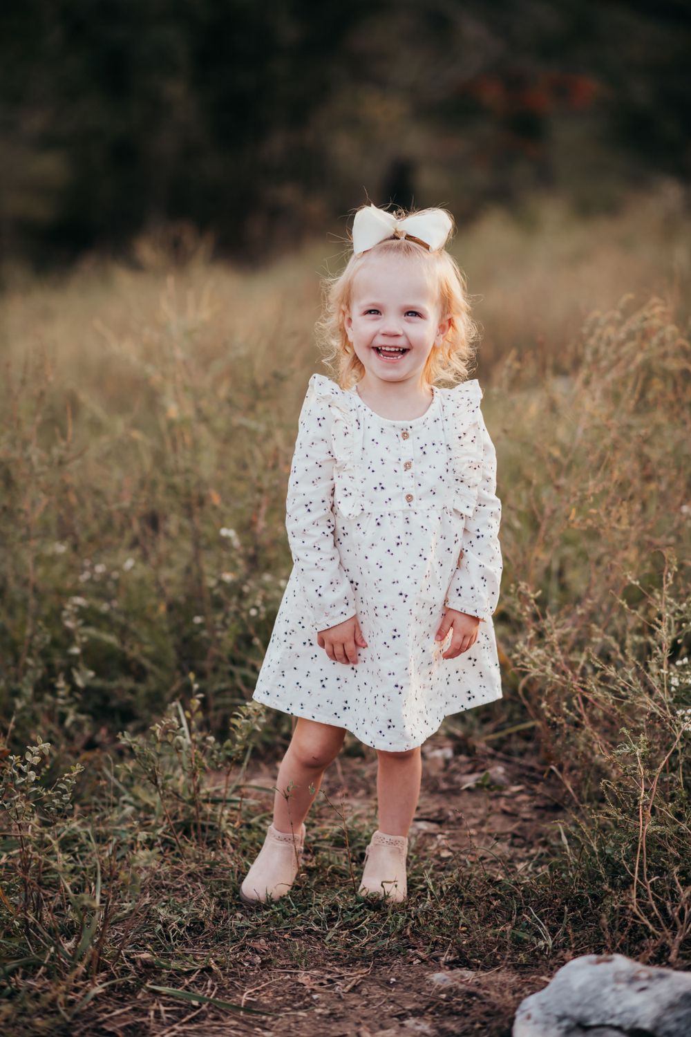 Joyful toddler in white floral dress standing on dirt path.