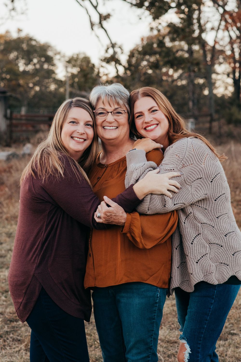 Three generations share a warm embrace while wearing cozy fall sweaters in a rustic outdoor setting at sunset.