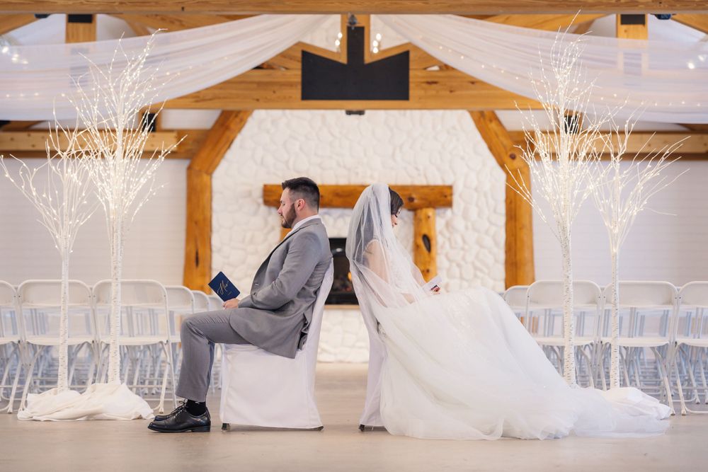Newlyweds sit back-to-back in a white ceremony space decorated with illuminated trees.