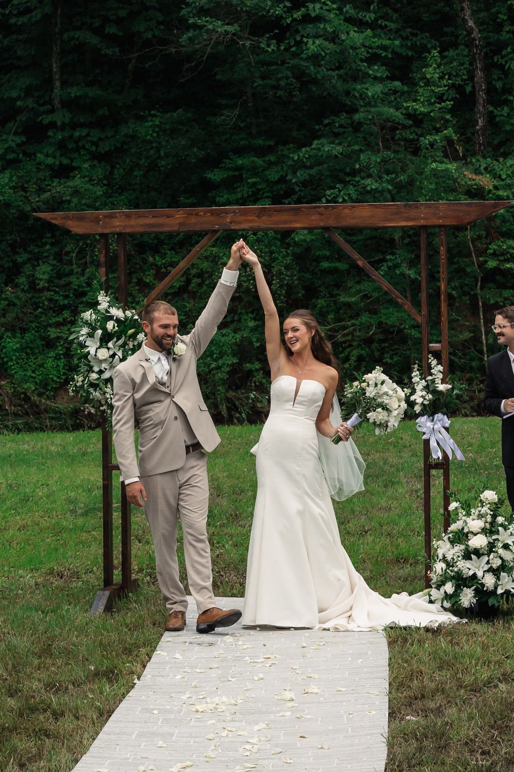 A joyous wedding ceremony under a rustic wooden arch decorated with white flowers in a lush green outdoor setting.