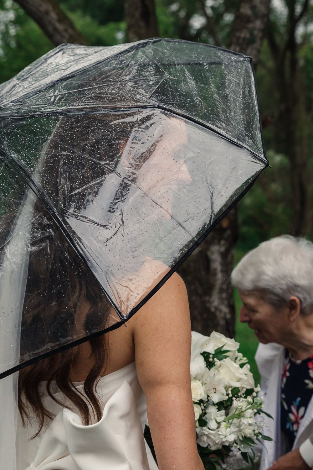 Clear umbrella protecting from rain during outdoor wedding with white floral bouquet.