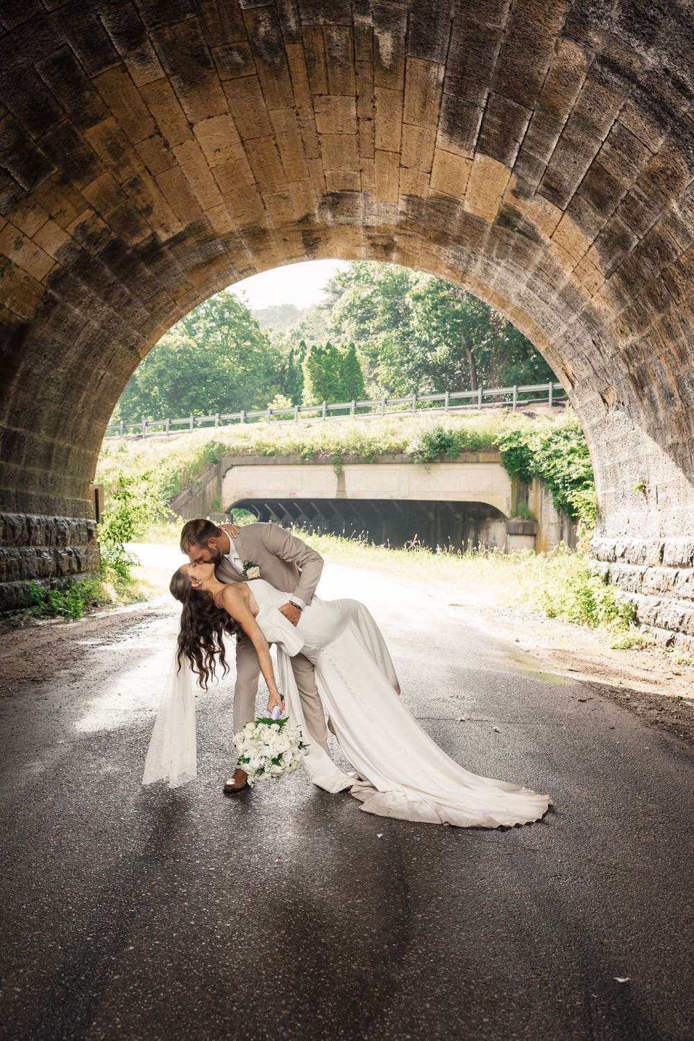 Romantic wedding photo captured under a stone bridge arch with flowing white dress.
