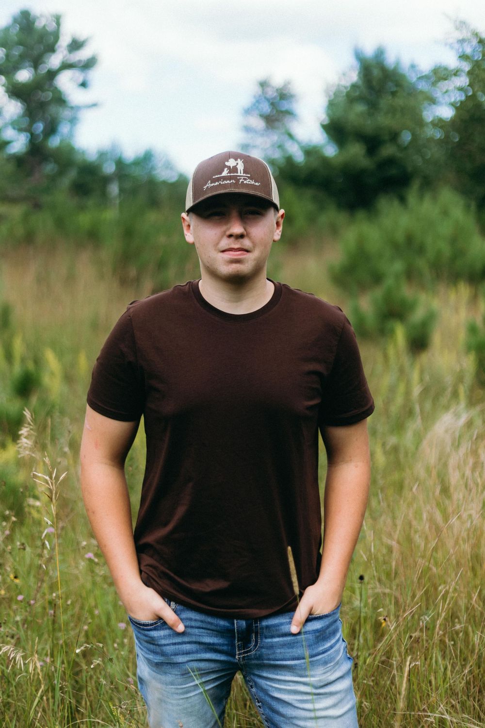 Person wearing a brown t-shirt and baseball cap stands in a field of tall grass on an overcast day.