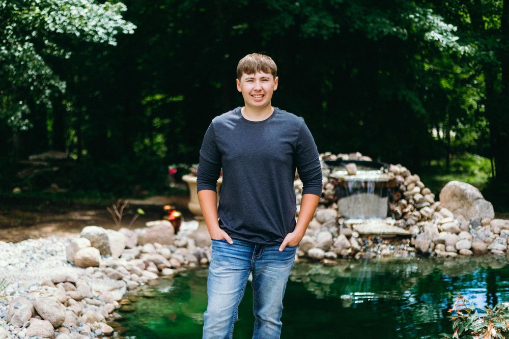 Man wearing a navy t-shirt and blue jeans stands near a pond surrounded by rocks and greenery.