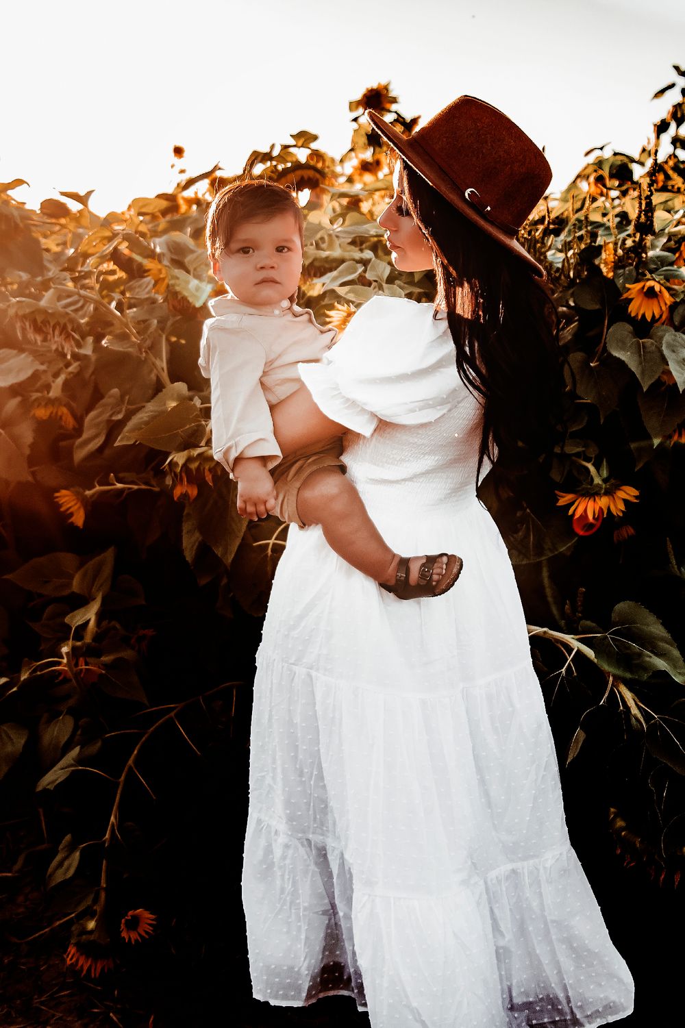 A small child in a white lacy dress stands in front of green foliage during golden hour sunset lighting.