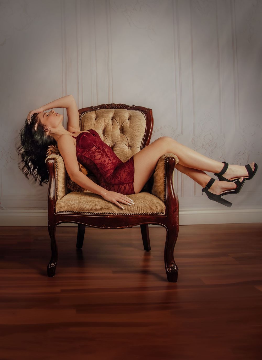 A figure in red lingerie reclines dramatically across a vintage beige chaise lounge against a plain wall.
