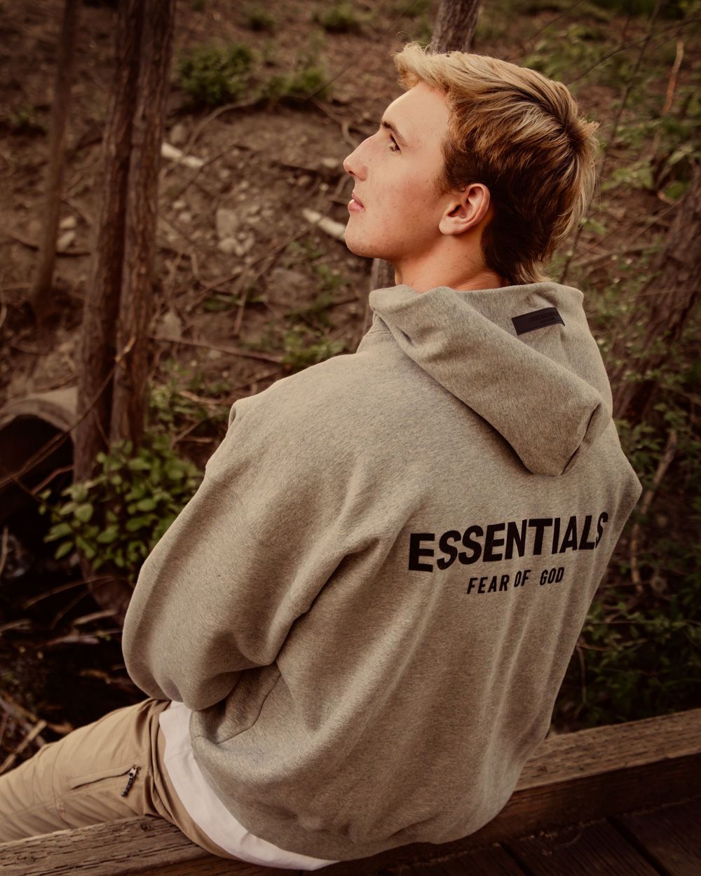 Person wearing a gray hoodie with "ESSENTIALS" text on the back sits outdoors in a wooded setting.