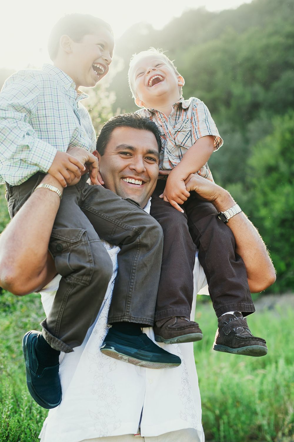 Happy father carrying two laughing young boys on his shoulders outdoors in a sunny green field.