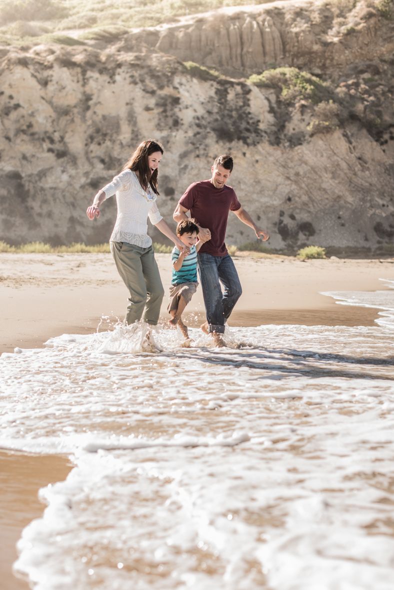 Family of three joyfully splashing in ocean waves on a sunny beach with sandy cliffs in the background.