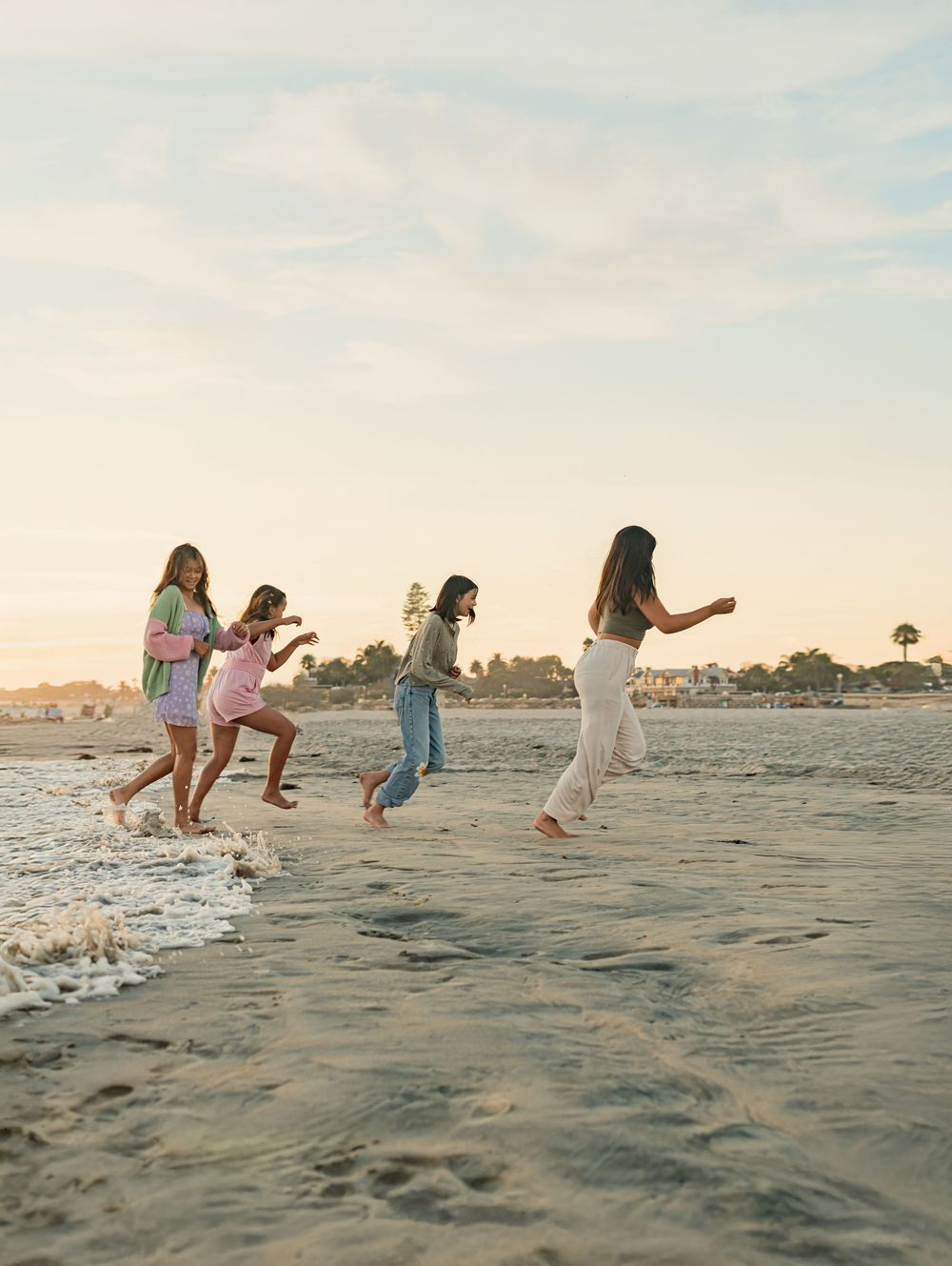 Four girls running and playing along a sandy beach at sunset with gentle waves and a coastal town in the background.