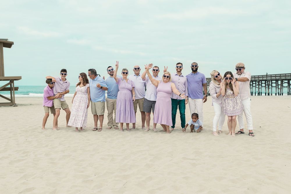 Large family group posing together on sandy beach near pier, wearing coordinated pastel outfits.
